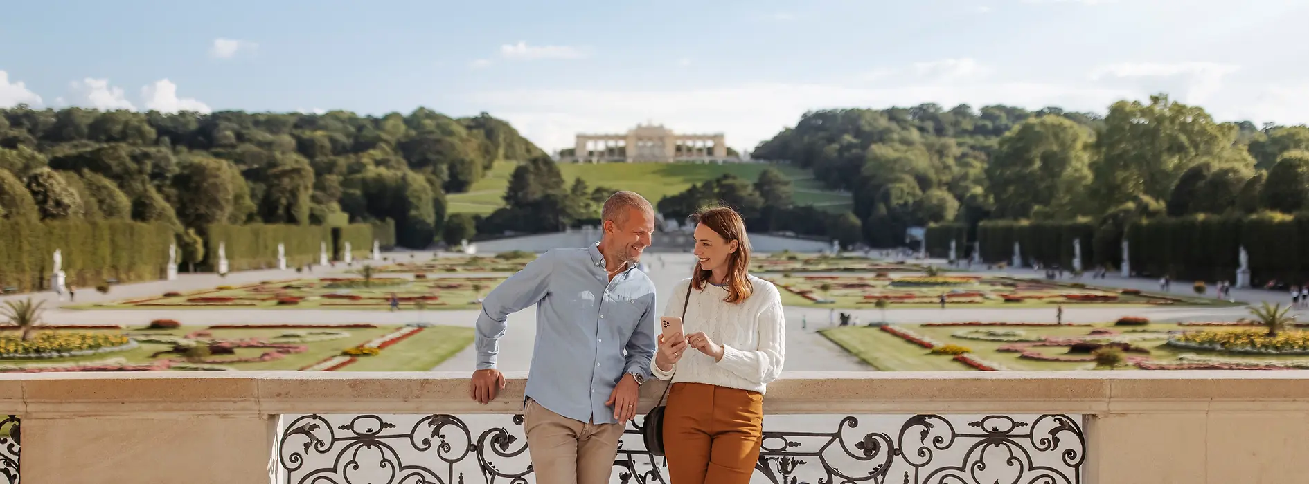 Un couple regarde son téléphone portable ensemble. En arrière-plan, les jardins du château de Schönbrunn et la Gloriette.