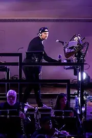 El dúo de música electrónica Camo & Krooked en el escenario con iluminación púrpura, con otros músicos tocando instrumentos clásicos frente a ellos